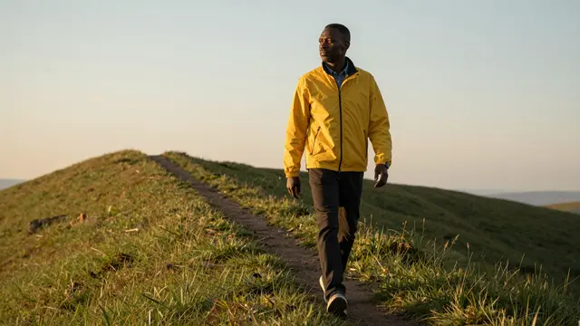 A Black man in a yellow jacket walks along a narrow grassy hill path at golden hour