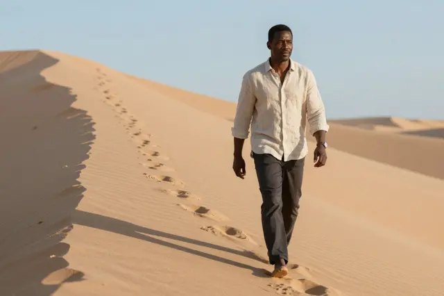 A Black man in a linen shirt walks barefoot along a sand dune ridge with footprints trailing behind him at sunrise.