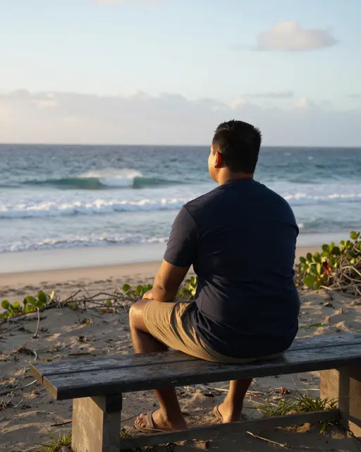 A man viewed from behind sits on a beach bench watching rolling ocean waves at sunrise.