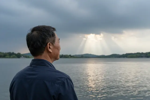 An East Asian man in his sixties stands looking toward sunbeams breaking through storm clouds over a lake.