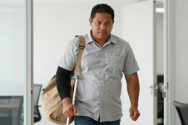 Pacific Islander man in his thirties walks through a bright office entrance using a forearm crutch with a canvas bag slung