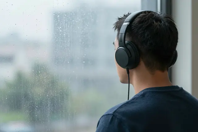 Viewed from behind, an adult man wearing plain black headphones looks out a rain-streaked window.