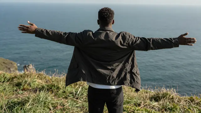 Back view of a Black man with outstretched arms standing on a grassy cliff overlooking the sea