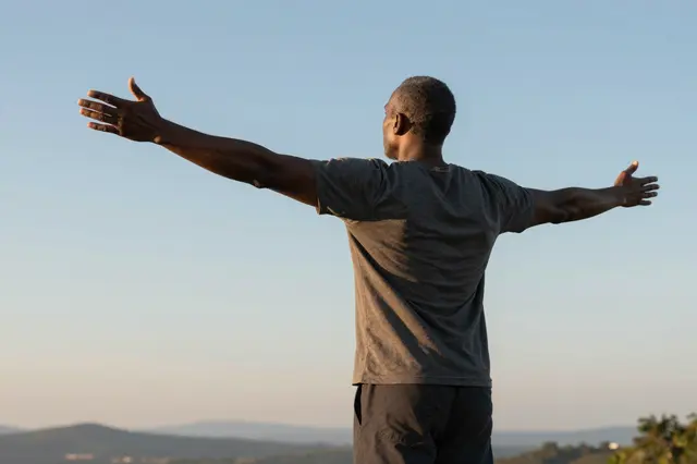 Rear view of a middle aged Black man standing on a hilltop with arms spread wide as the sun rises
