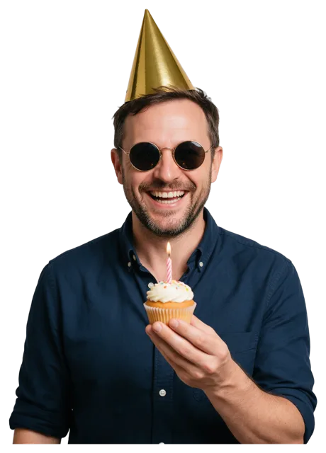 A smiling man in a gold party hat and sunglasses holds a cupcake with a lit candle.