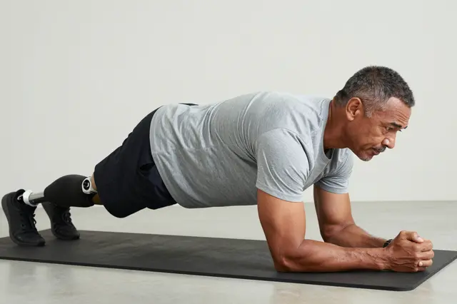 Middle aged man with a prosthetic leg holds a forearm plank on a black exercise mat in a plain indoor space