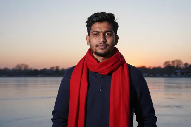 South Asian man in a red scarf stands beside a frozen lake at soft sunset
