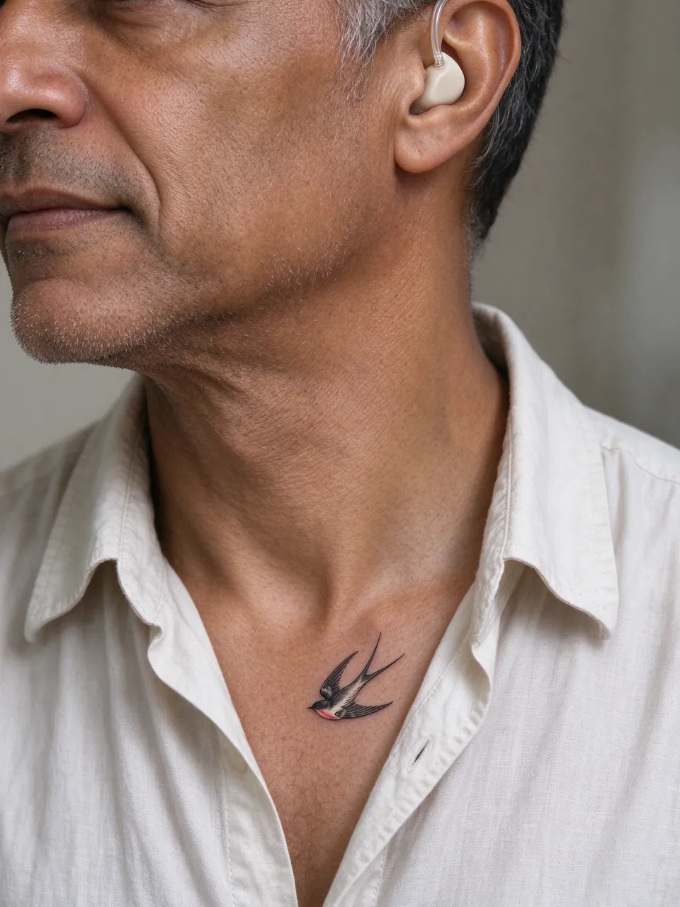 Close-up of a mixed-race man in his fifties showing a collarbone swallow tattoo and in-ear hearing aid