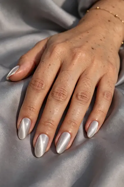 A hand with glossy silver almond nails rests smoothly draped grey silk fabric.