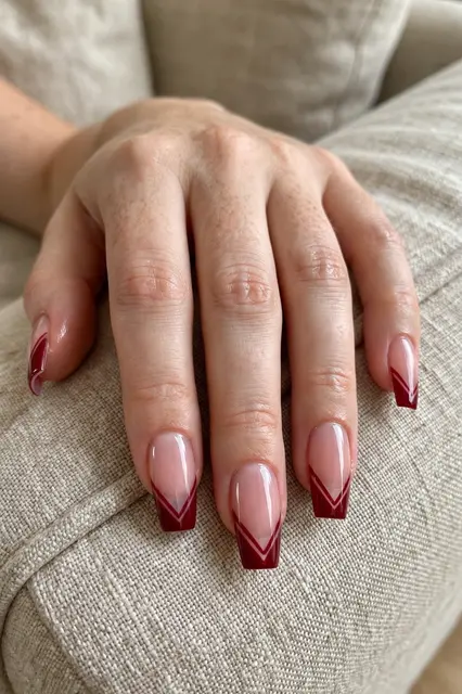A close-up of a woman's hand with deep red chevron French tip coffin nails resting on a beige linen sofa arm