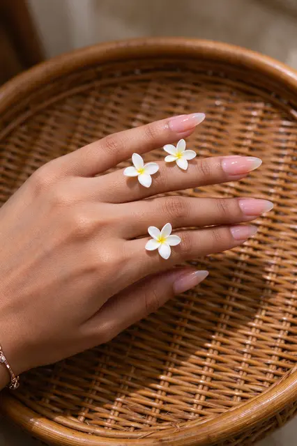 A South Asian woman's hand with almond sheer pink nails rests on a rattan tray with small plumeria flowers between fingers