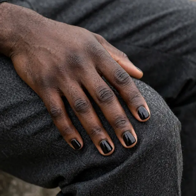 Close-up of a Black man's hand with glossy black square nail polish resting on charcoal wool trousers