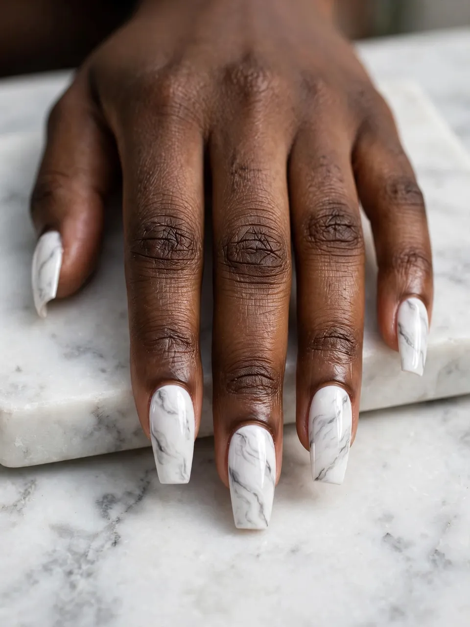 A Black woman's hand with marble-patterned coffin nails rests on a white marble slab