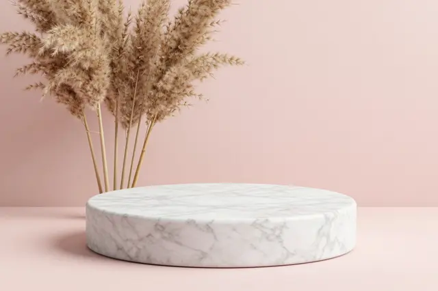 A white veined marble display podium sits next to dried pampas grass against a soft blush pink backdrop