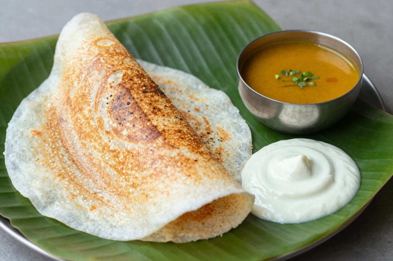 Crispy masala dosa served with sambar and coconut chutney on a fresh green banana leaf plate.