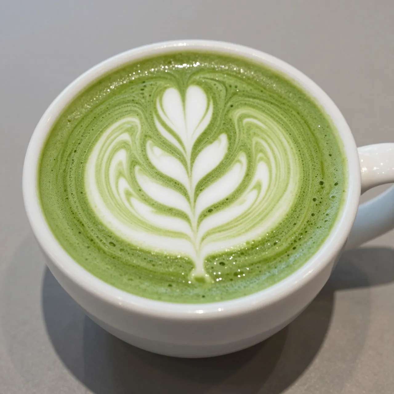 Top-down view of a white ceramic cup holding a matcha latte with intricate leaf latte art on a gray table surface.