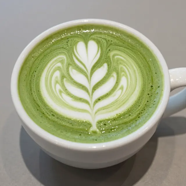 Top-down view of a white ceramic cup holding a matcha latte with intricate leaf latte art on a gray table surface.