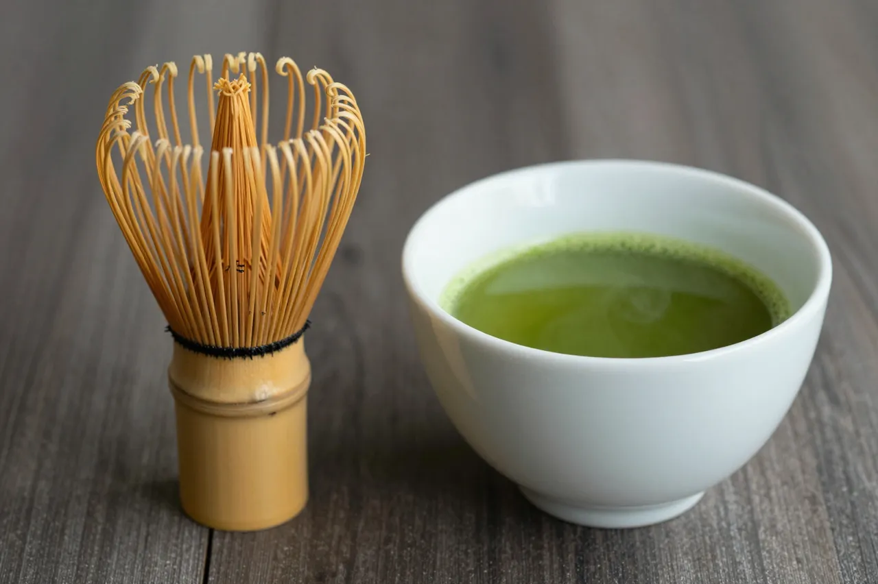 A bamboo matcha whisk sits next to a white ceramic bowl of prepared green matcha on a dark wooden table.