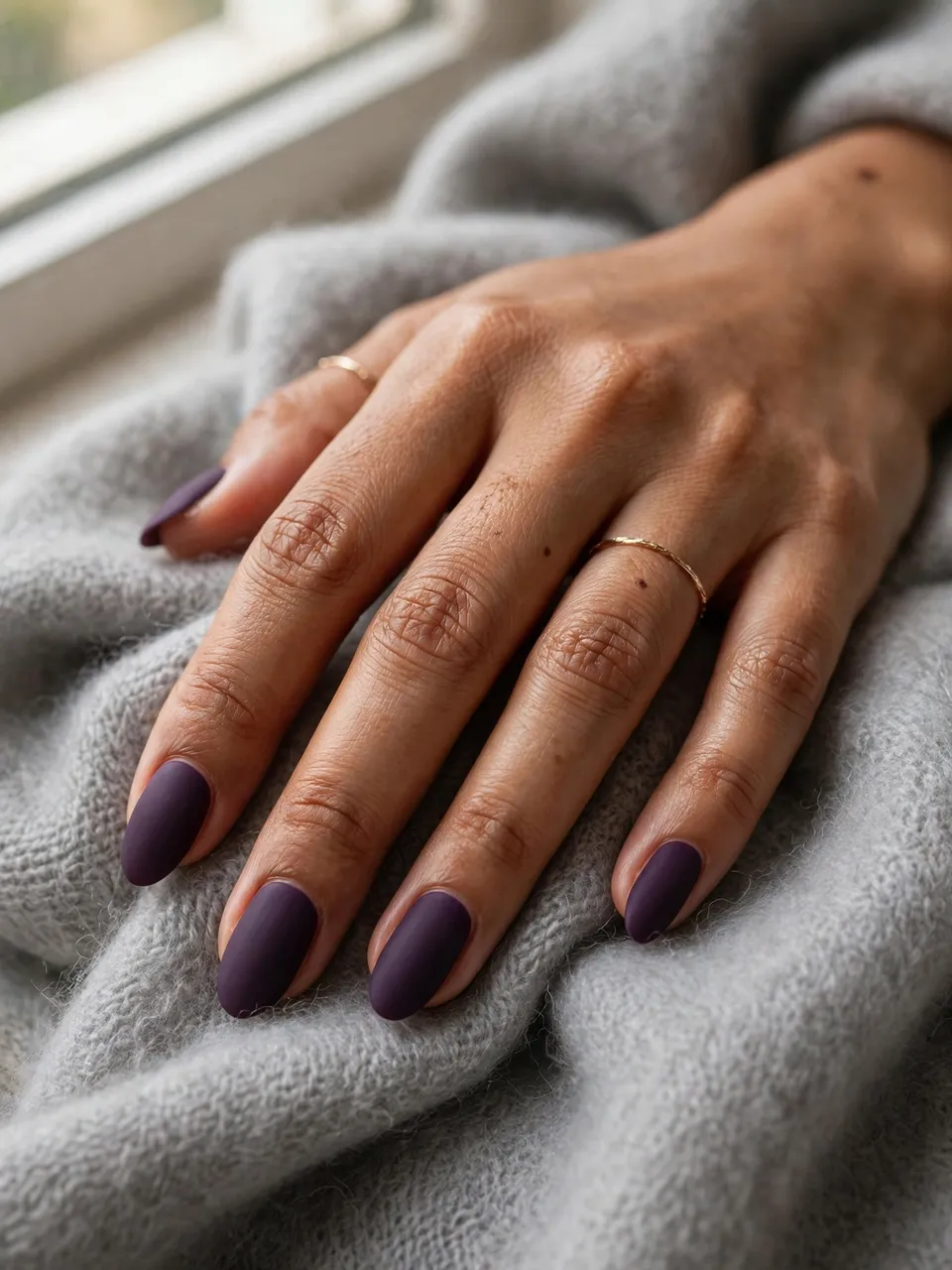 A Southeast Asian woman's hand with matte deep purple almond nails rests on soft grey cashmere near a sunlit window