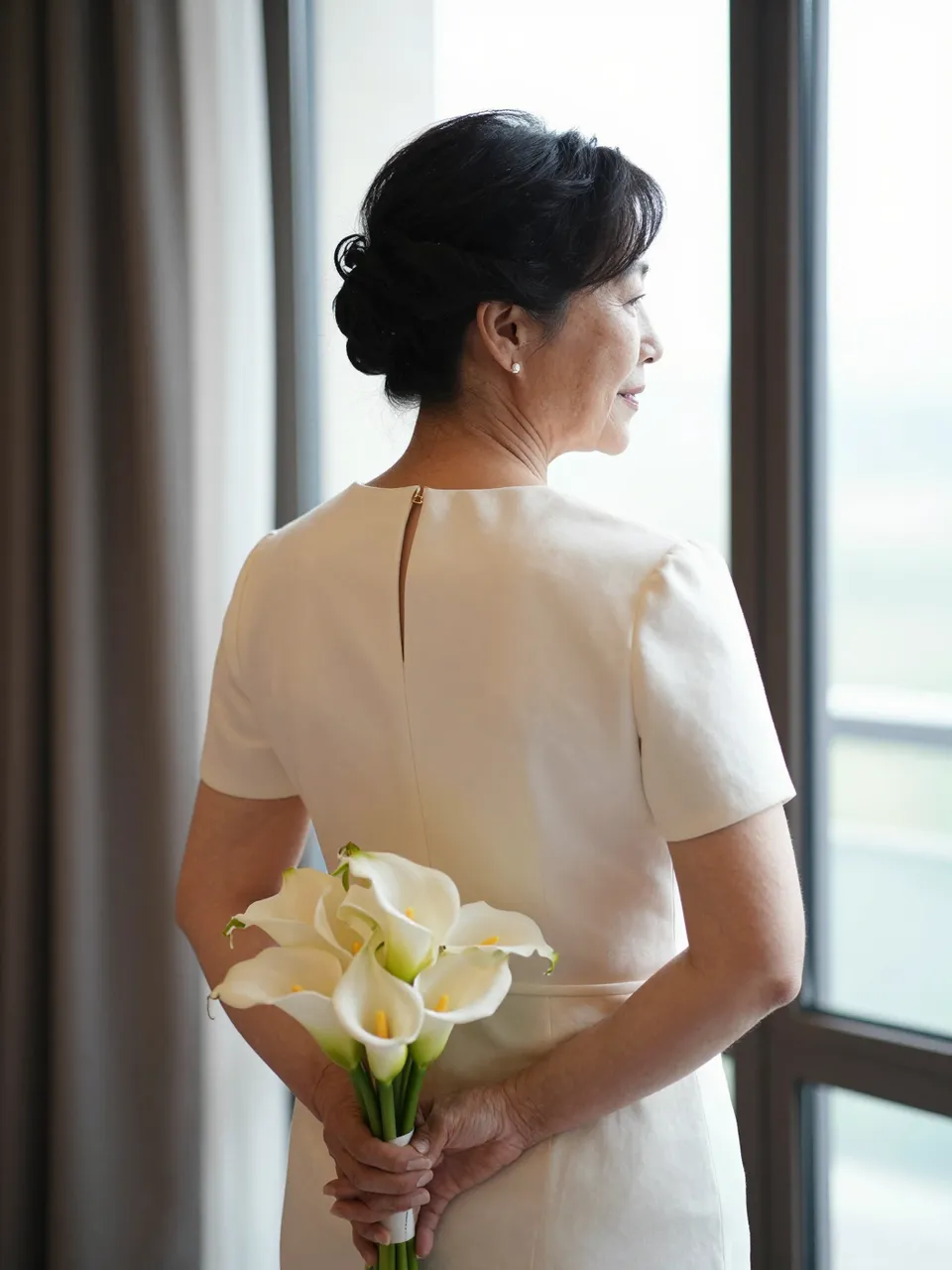 Back view of an older East Asian bride holding a calla lily bouquet behind her back while looking out a window