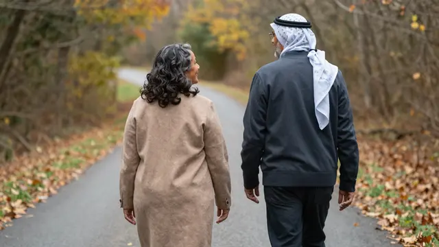 Two senior adults walk side by side along a leaf-strewn autumn paved road viewed from behind
