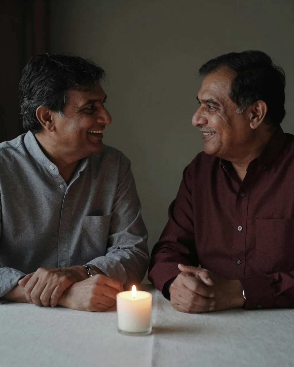 Two smiling older South Asian men in their sixties look at each other across a lit candle on a linen table