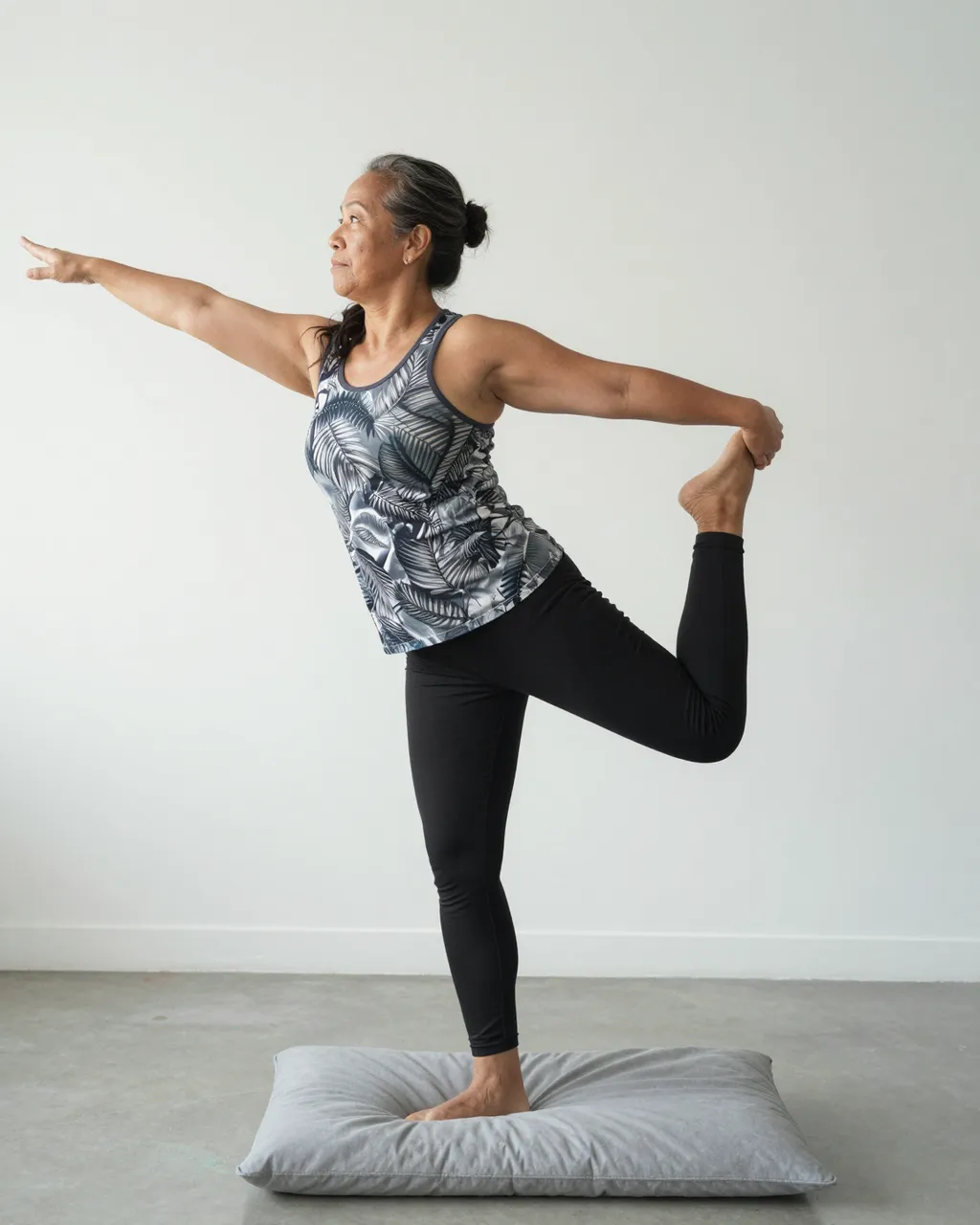 A mature Pacific Islander woman holds a standing yoga balance pose on a cushion in a bright studio