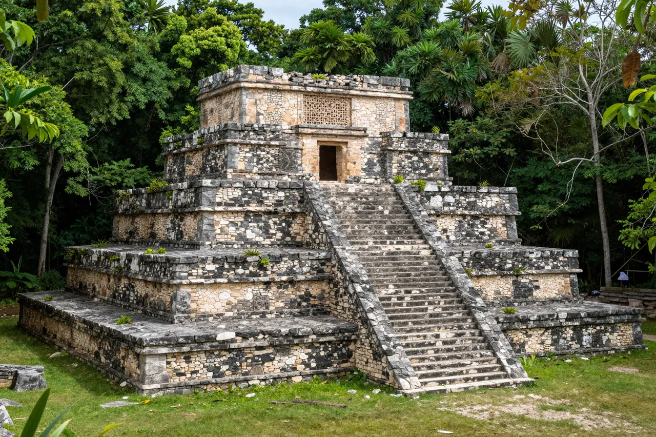 An ancient tiered stone Mayan ruin with a central staircase sits surrounded by dense tropical jungle
