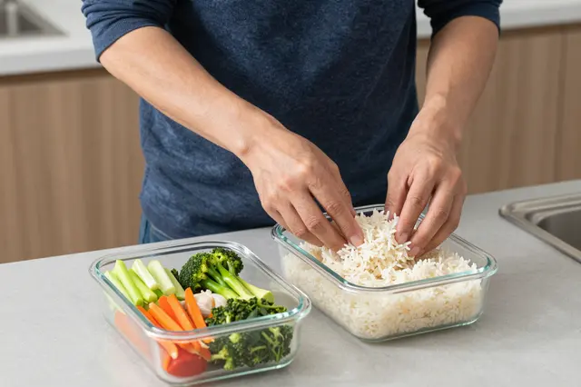 An adult man portions cooked rice into a glass meal prep container next to fresh cut vegetables on a kitchen counter.