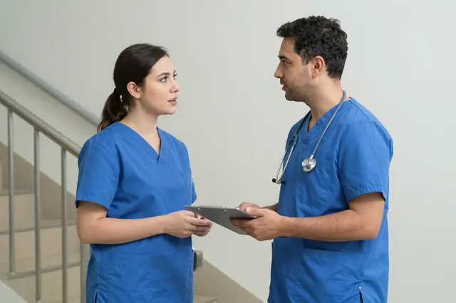 Two doctors in blue scrubs converse while standing in a hospital stairwell holding a clipboard