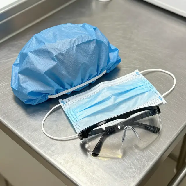 Disposable blue surgical cap, face mask and safety glasses laid flat on a brushed stainless steel counter, top down view.