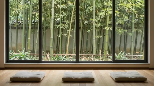 Three floor meditation cushions sit on wooden flooring in front of a large window overlooking a lush bamboo garden.