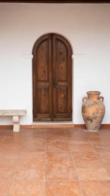 Arched wooden entry door flanked by a stone bench and clay urn above terracotta tile flooring