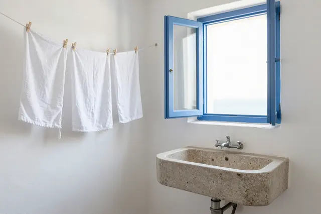 White linen towels dry on a line next to a stone sink beneath an open blue-framed window