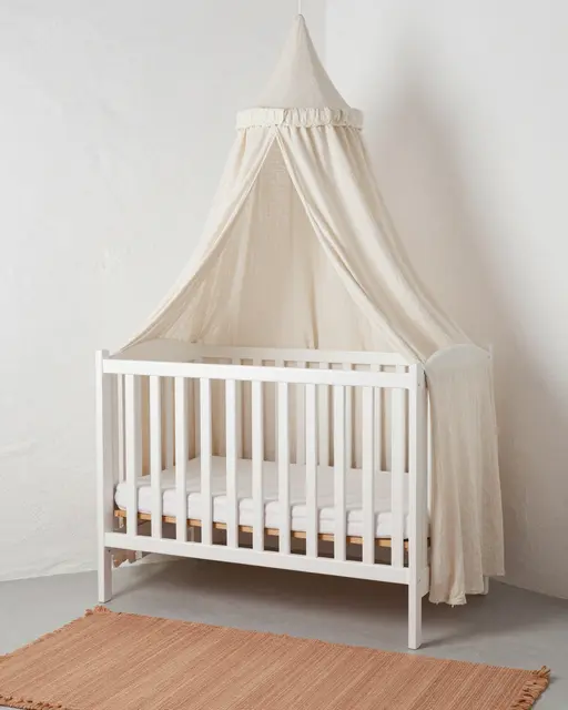 Whitewashed wooden crib with linen canopy sits above a terracotta rug against plaster walls