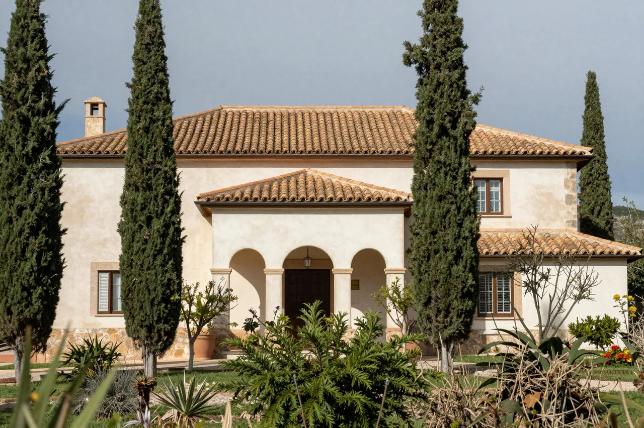 Stucco mediterranean villa with terracotta roof framed by tall cypress trees and garden foliage under an overcast sky