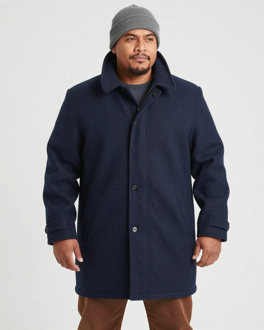 Pacific Islander man in his thirties wearing a navy wool coat, gray beanie and brown trousers stands against a plain light