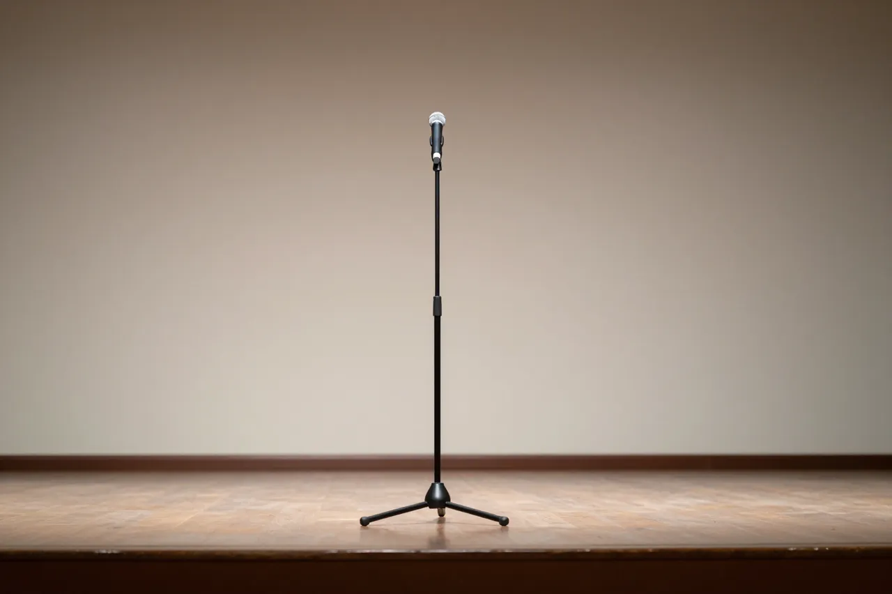 A single microphone on a stand sits centered on an empty wooden stage against a plain lit wall