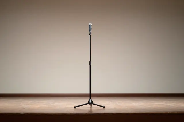 A single microphone on a stand sits centered on an empty wooden stage against a plain lit wall
