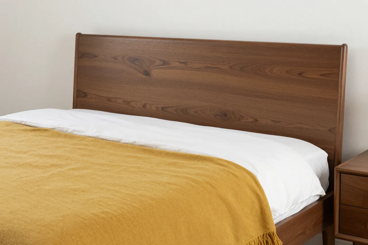 A walnut mid-century bed with white linens and a mustard throw sits beside a matching nightstand against a plain wall