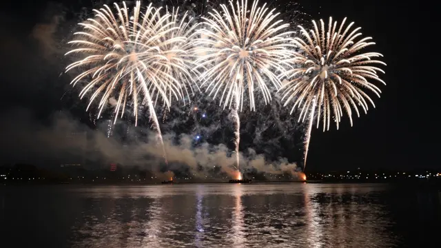 Bright golden fireworks burst over a calm lake with light reflections on the dark water at night