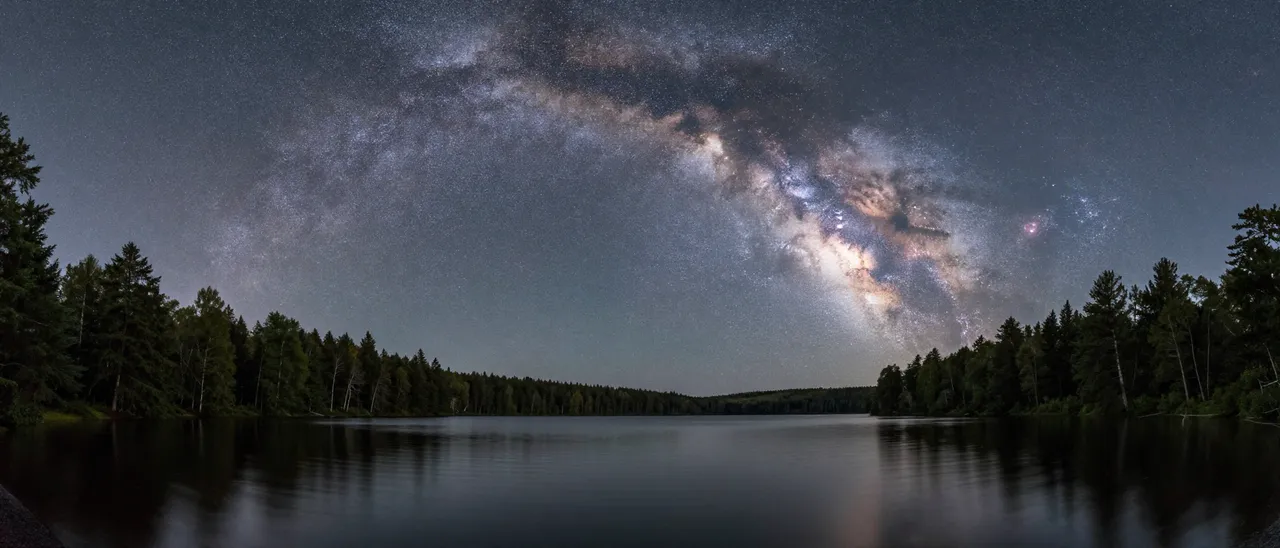 Panoramic arch of the milky way stretches across the night sky above a calm forested lake.
