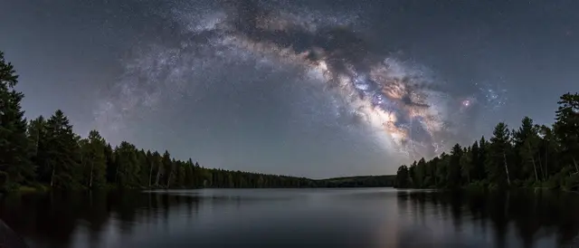 Panoramic arch of the milky way stretches across the night sky above a calm forested lake.