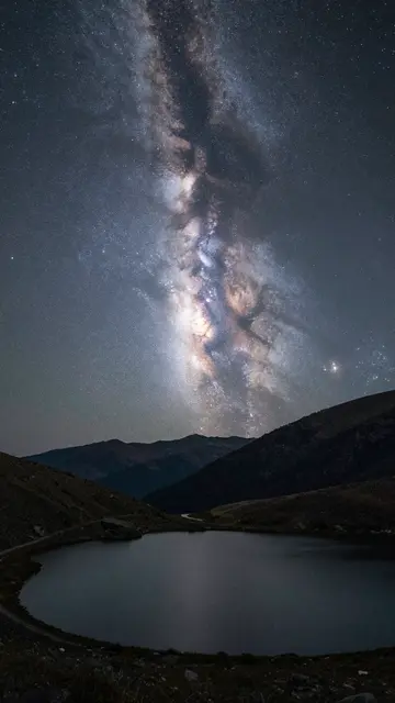 Bright milky way core rises above calm mountain lake and dark alpine ridgelines at night
