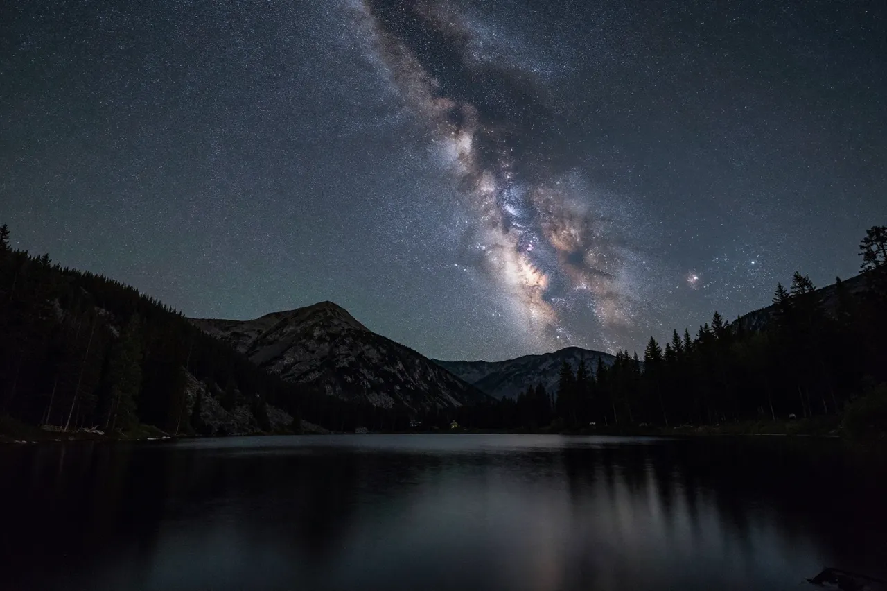 Bright Milky Way band stretches across the night sky above a calm mountain lake and forested peaks