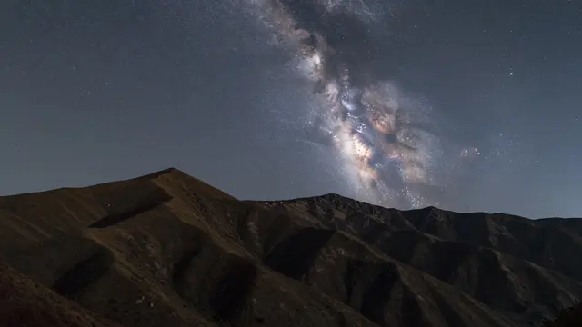 Bright Milky Way galaxy stretches across a dark night sky above silhouetted arid mountain ridges