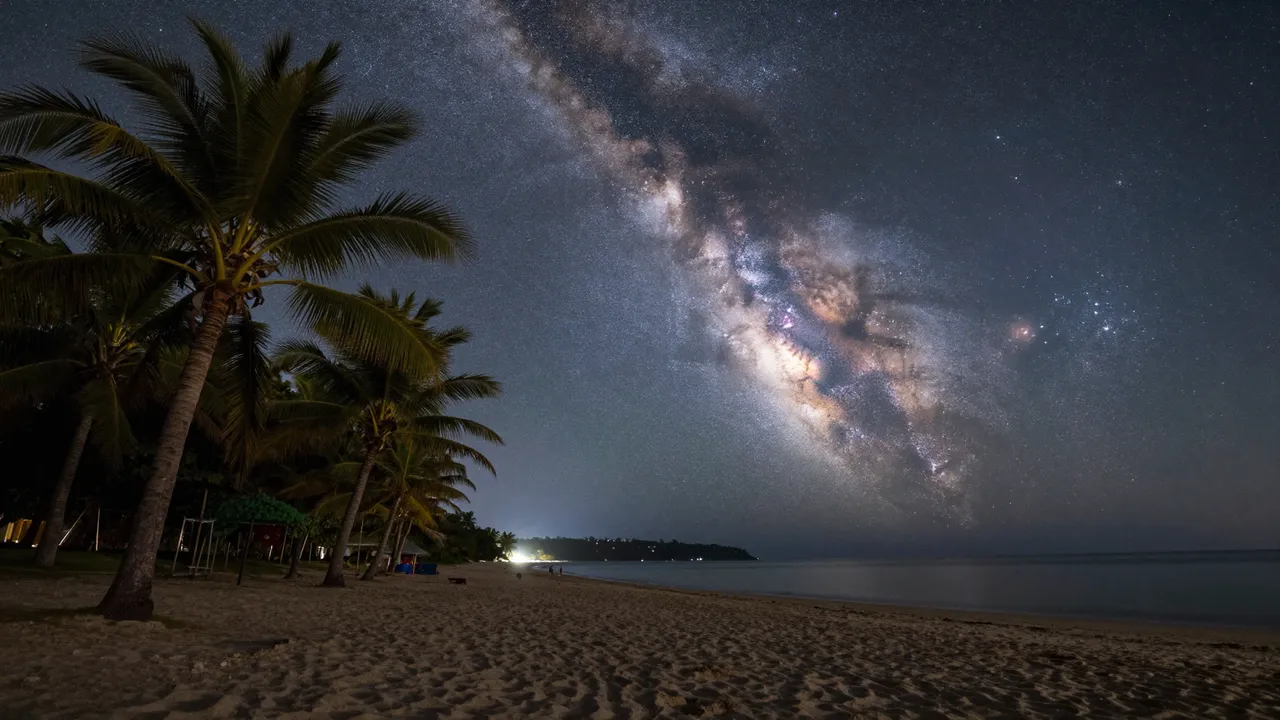 Bright milky way stretches across the night sky above a sandy palm-lined ocean beach