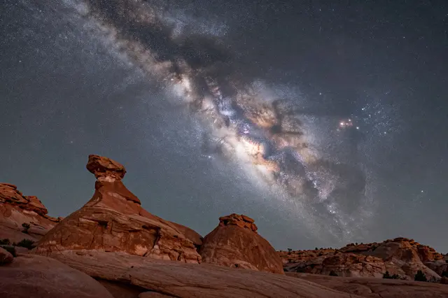 The glowing milky way core arcs across a dark night sky above eroded red rock hoodoos.