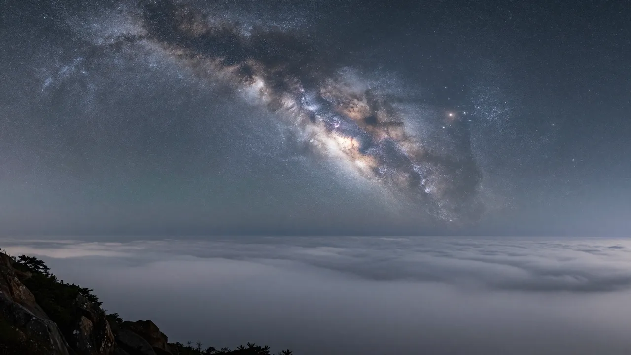 Bright arching milky way galaxy stretches over a calm sea of clouds viewed from a rocky mountain summit at night