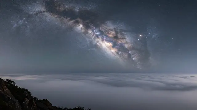 Bright arching milky way galaxy stretches over a calm sea of clouds viewed from a rocky mountain summit at night